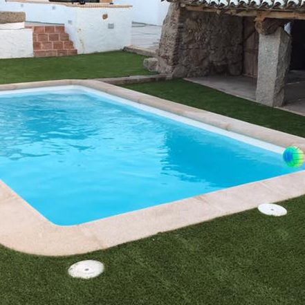 Piscina de agua clara rodeada de césped artificial y una pared de piedra.