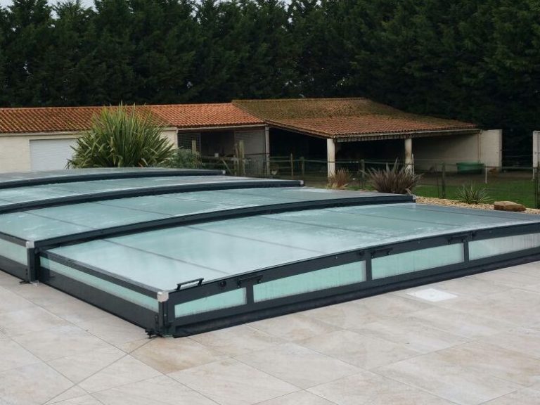 Cubierta de piscina de policarbonato con estructura metálica, rodeada de jardín y una casa al fondo.